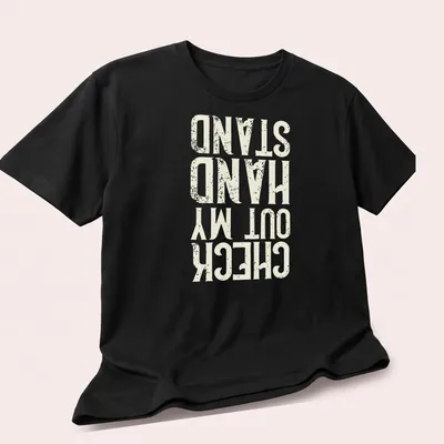 Distressed typography t-shirt featuring the phrase Check Out My Hand Stand in white, cream, and gold. — printed on a unisex t-shirt (angled view)