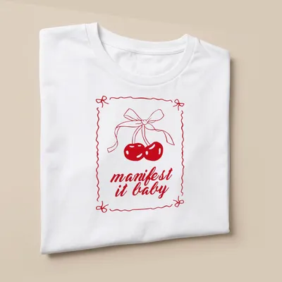 Kawaii line-art shirt with cherries and a bow, featuring "manifest it baby" text — printed on a unisex t-shirt (angled view)