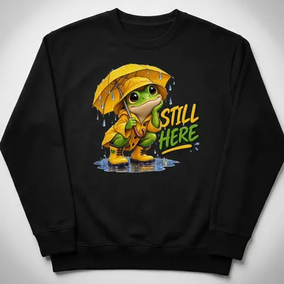 Colorful cartoon frog in a yellow raincoat with an umbrella and text 'STILL HERE' — printed on a unisex sweatshirt