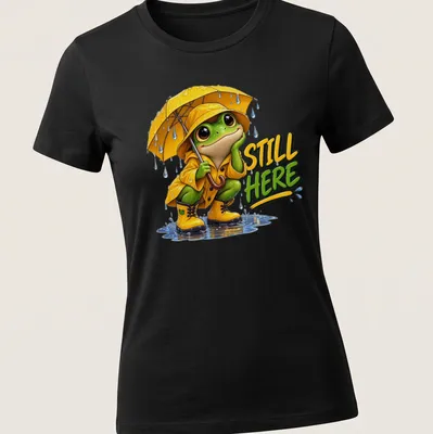 Colorful cartoon frog in a yellow raincoat with an umbrella and text 'STILL HERE' — printed on a women's shirt