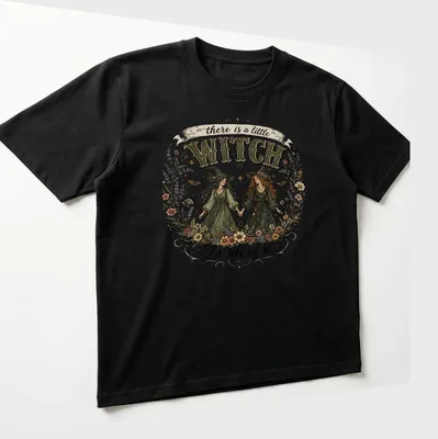Two whimsical witches with the text 'there is a little WITCH In all of us' in vintage style — printed on a unisex t-shirt (angled view)
